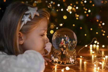 Photo for Girl looking at a glass ball with a scene of the birth of Jesus Christ near christmas tree - Royalty Free Image