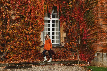 Foto de Mujer en un suéter naranja cerca de una pared con uvas silvestres en otoño - Imagen libre de derechos