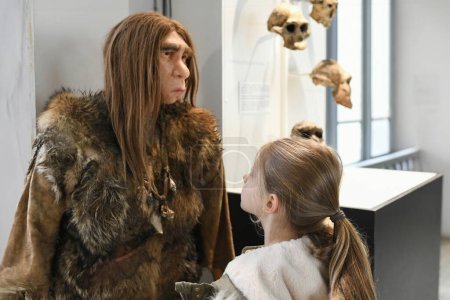 The girl looks at wax figure of a Neanderthal with long hair in animal skin
