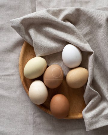 Foto de Nature colors Easter eggs. Eggs on a wooden plate on a beige napkin. A chicken egg as a valuable nutritious product. Top view flat lay - Imagen libre de derechos