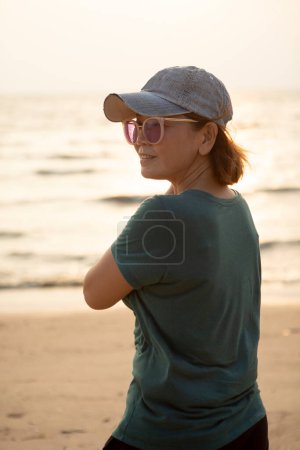 Téléchargez les photos : Portrait de femme asiatique portant un bonnet de jeans debout à la plage de la mer contre la belle lumière du coucher du soleil - en image libre de droit