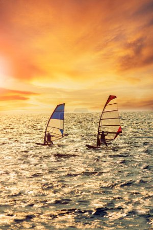 Téléchargez les photos : Deux hommes jouant à la planche à voile sur la mer se précipitant contre l'éclairage magnifique coucher de soleil - en image libre de droit