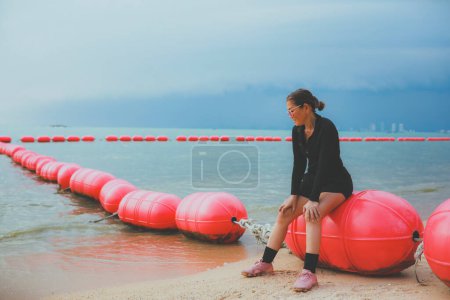 Photo for Woman wearing black  running sport suit sitting on floating barrier on sea beach - Royalty Free Image