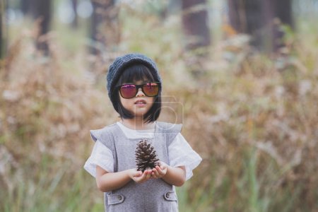 Téléchargez les photos : Petite fille portant capuche en laine et lunettes de soleil tenant des cônes de pin sauvage à la main - en image libre de droit