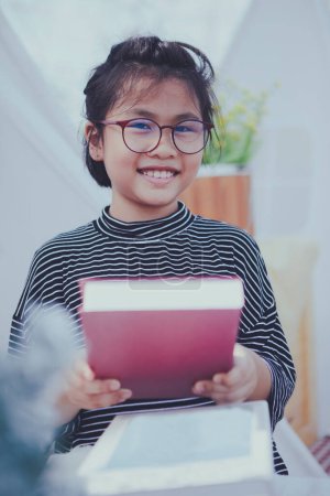 Téléchargez les photos : Asiatique fille tenant Gros école livre dans la main et toothy sourire avec bonheur - en image libre de droit