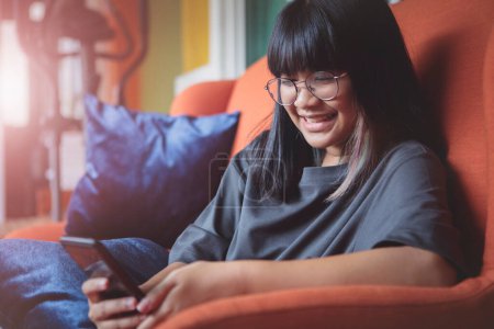 Téléchargez les photos : Asiatique adolescent lecture message dans smartphone avec bonheur visage à la maison salon - en image libre de droit