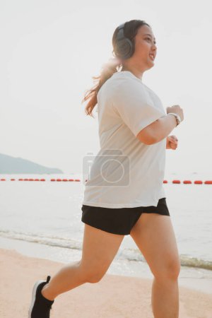Téléchargez les photos : Femme potelée portant des vêtements de sport et haedphone courir sur la plage de la mer avec bonheur - en image libre de droit