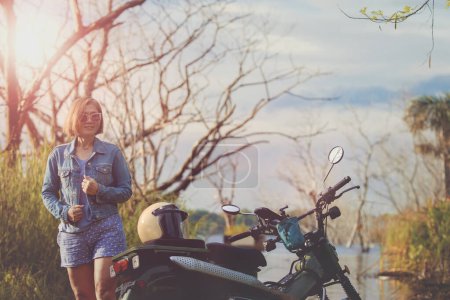 Téléchargez les photos : Belle femme dents souriant avec bonheur debout à côté de la petite moto enduro au champ de jungle - en image libre de droit