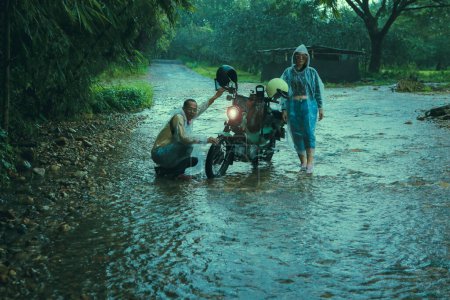 Téléchargez les photos : Couples de motards asiatiques portant des vêtements de pluie en plastique entretien petite moto enduro dans le ruisseau peu profond parmi la pluie tombant - en image libre de droit