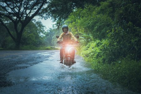 Téléchargez les photos : Homme chevauchant petite moto endurom traversant ruisseau peu profond parmi la pluie tombant à la forêt - en image libre de droit
