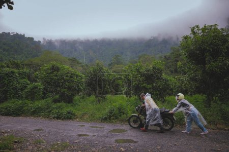 Téléchargez les photos : Couples asiatiques collaboré pour pousser une moto défectueuse au milieu de la route forestière - en image libre de droit