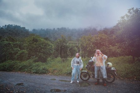 Téléchargez les photos : Couples de motards asiatiques vêtus d'imperméable en plastique tachant à côté de la petite moto enduro contre les nuages pluvieux sur piste de pays thailand - en image libre de droit