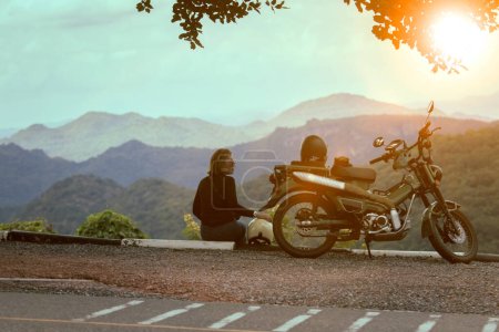Téléchargez les photos : Femme assise à côté de la moto à beau point de vue et regardant la scène du coucher du soleil - en image libre de droit