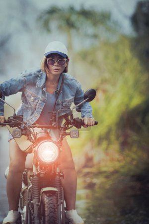 Téléchargez les photos : Belle femme portant une veste en jean bleu assis sur moto enduro sur fond naturel coloré - en image libre de droit