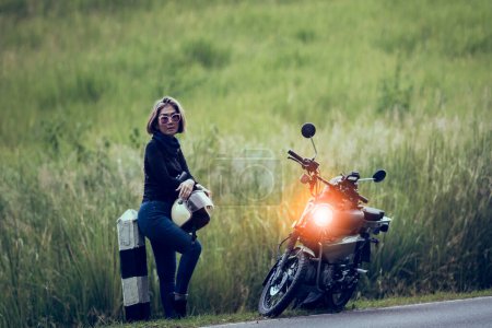 Téléchargez les photos : Jolie femme tenant casque de sécurité debout à côté de la petite moto enduro contre champ d'herbe verte - en image libre de droit