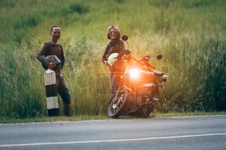 Téléchargez les photos : Asiatique couple tenant casque de sécurité debout à côté de petite moto enduro contre belle herbe verte - en image libre de droit