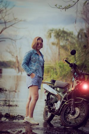 Téléchargez les photos : Belle femme portant une veste en jean bleu debout à côté de la moto enduro sur fond naturel coloré - en image libre de droit