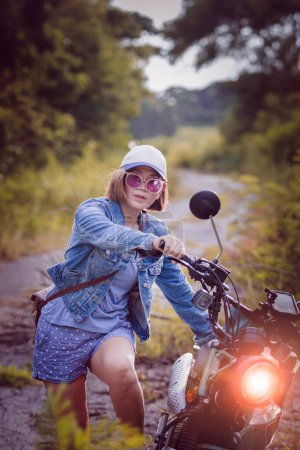Téléchargez les photos : Belle femme portant une veste en jean bleu assis sur moto enduro sur fond naturel coloré - en image libre de droit