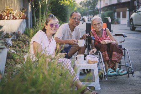 Téléchargez les photos : Asiatique famille avec grand-mère assis sur fauteuil roulant profiter matin café à la maison village - en image libre de droit