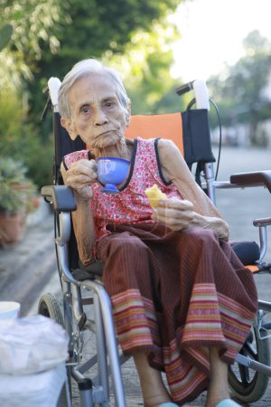 Téléchargez les photos : Asiatique vieille femme assis sur whell chaise et boire boisson chaude - en image libre de droit
