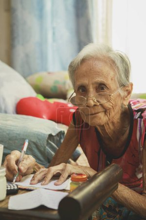 Photo for Old asian woman writing wording on white paper at home living room - Royalty Free Image