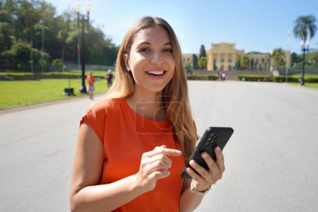 Téléchargez les photos : Happy beautiful young woman looking at camera and holding mobile phone outdoor with blurred city park on background. City lifestyle people technology. - en image libre de droit