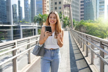 Photo for Young casual Brazilian woman watching her mobile phone walking in the city - Royalty Free Image