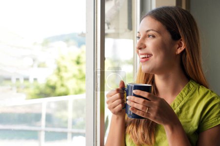 Foto de Empresaria sosteniendo una taza de café cerca de la ventana de su oficina en la mañana temprano. Concepto café y trabajo. - Imagen libre de derechos