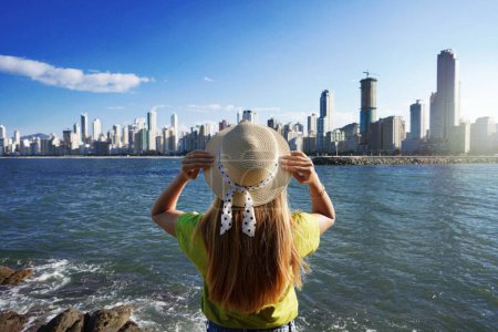 Photo for Overbuilding on the coast. Young woman with hat looking to skyline full of skyscrapers on south coast of Brazil. - Royalty Free Image