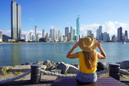 Urlaub im Balneario Camboriu, Brasilien. Rückansicht einer jungen Frau mit Hut, die entspannt auf einer Bank sitzt und die Skyline von Balneario Camboriu, Santa Catarina, Brasilien genießt.