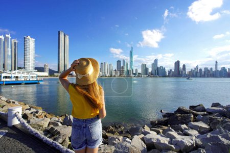 Rückansicht einer jungen Frau mit Strohhut, die entspannt den Panoramablick auf die Skyline von Balneario Camboriu, Santa Catarina, Brasilien genießt
