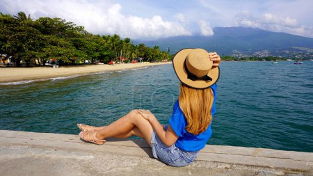 Schöne junge Frau mit Hut sitzt auf einem Steg und blickt auf den Panoramablick auf die Insel Ilhabela, Brasilien