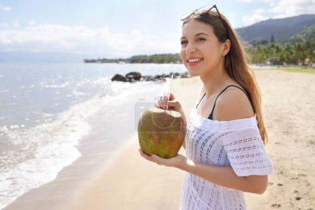 Mujer joven sosteniendo un coco verde fresco disfrutando de beber en la playa