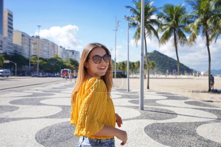 Téléchargez les photos : Portrait de voyageuse détendue souriante marchant le long de la promenade de la plage de Copacabana et se tournant vers la caméra, Rio de Janeiro, Brésil - en image libre de droit