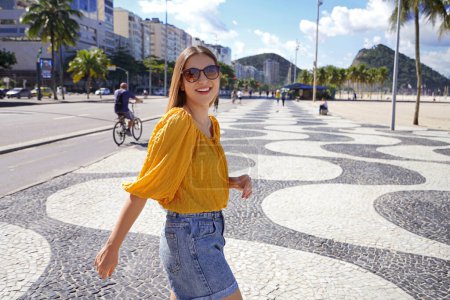 Téléchargez les photos : Heureuse femme souriante marchant le long de la promenade de la plage de Copacabana et se tourne vers la caméra, Rio de Janeiro, Brésil - en image libre de droit