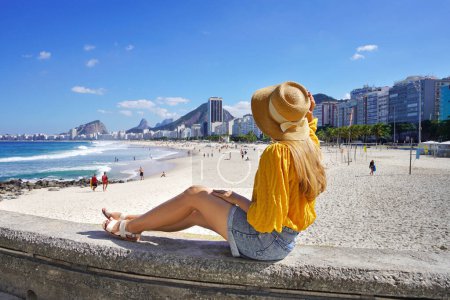 Ferien in Rio de Janeiro. Rückansicht der schönen Mode Mädchen sitzt an der Wand und genießt den Blick auf den Strand von Copacabana. Sommerurlaub nach Brasilien.