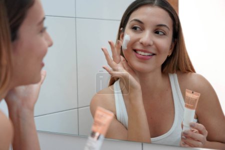 Daily routine. Smiling young woman looking at mirror using face cream.