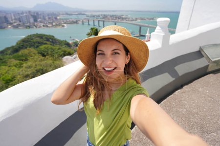 Foto de Mujer sonriente de moda viajera toma autorretrato con la vista panorámica de la ciudad de Vitoria en el estado de Espirito Santo, Brasil - Imagen libre de derechos