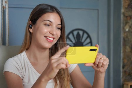 Brazilian girl with earbuds watching broadcasting on video sharing platform sitting on sofa in her living room. Woman video calls with her mobile phone at home.