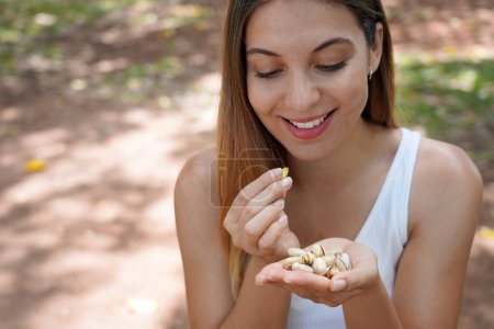 Téléchargez les photos : Belle femme saine mangeant des graines de pistache en plein air. Regarde les pistaches dans sa main. - en image libre de droit