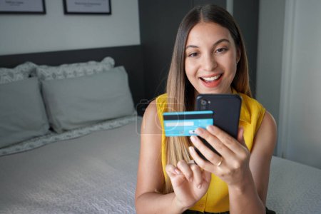 Paiement réussi. Joyeuse jeune femme excitée faisant des transactions en ligne avec son smartphone et sa carte de crédit confortablement à la maison. Achats en ligne et concept de banque en ligne.