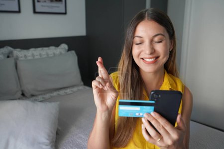 Joven mujer bonita esperanzada mantiene los dedos cruzados expresa un deseo sostiene smartphone y tarjeta de crédito espera sueños hechos realidad