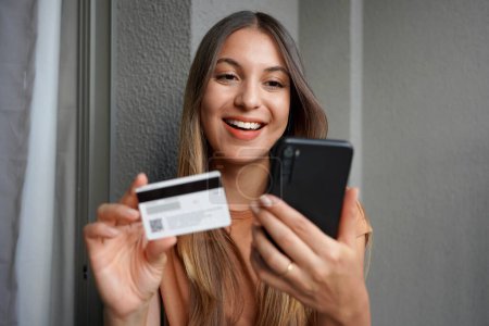 Primer plano de atractiva chica hispana hace compra en smartphone con tarjeta de crédito