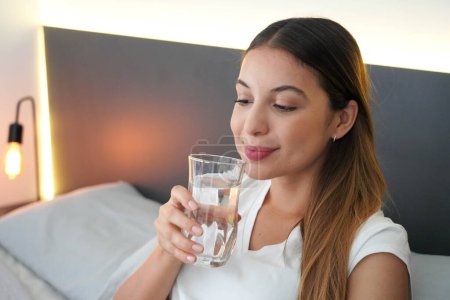 Schöne Mädchen im Bett trinkt ein Glas Wasser, das gerade am Morgen aufgewacht ist. Bedeutung einer guten täglichen Flüssigkeitszufuhr.