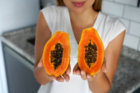 Unidentified young woman showing a papaya cut into two halves in the kitchen