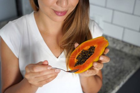Unidentified young woman holding half papaya fruit and spoon in the kitchen