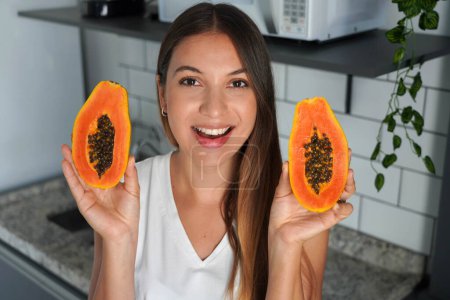 Hermosa joven que muestra una papaya cortada en dos mitades en la cocina