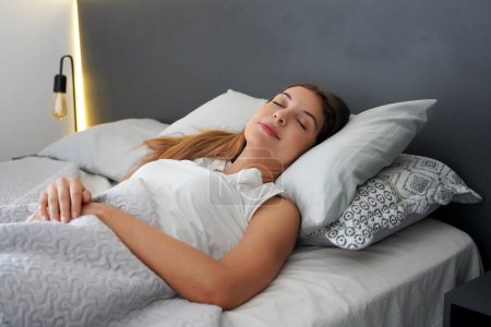 Young woman sleeping well in bed with soft pillows. Teenage girl has good night sleep. Lady enjoys fresh soft bedding linen and mattress in bedroom.