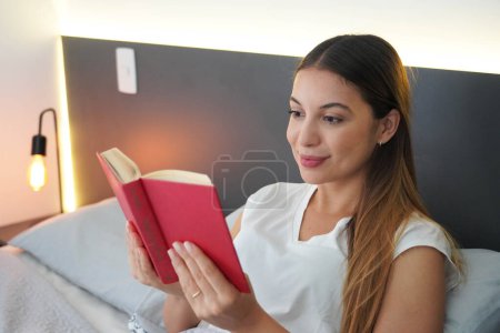 Nahaufnahme einer jungen Frau, die vor dem Schlafen im Bett unter den Decken ein Buch liest