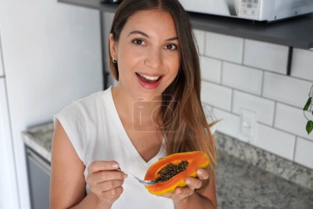 Retrato de una hermosa mujer sosteniendo media fruta de papaya y cuchara en la cocina. Mira la cámara..